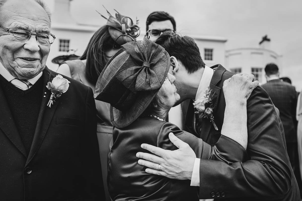 black and white photograph of the groom kissing his mother whilst his father grins at them behind