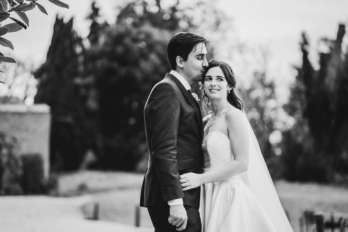 Black and white portrait of the bride and groom in the gardens of their wedding venue. the groom kisses his wife's forehead