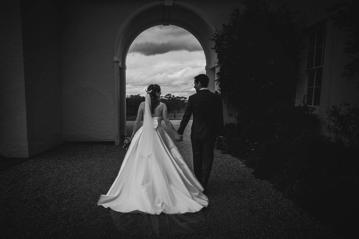the bride and groom walk away from the photographer under the arch at the Exeter wedding venue. the bride's dress train and veil flow behind her as she walks away