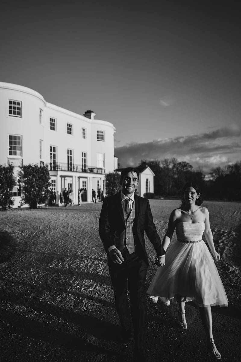 black and white photograph of side light hitting the newly-weds as they walk away from rockbeare Manor House in the background. wedding guests can be seen enjoying themselves behind