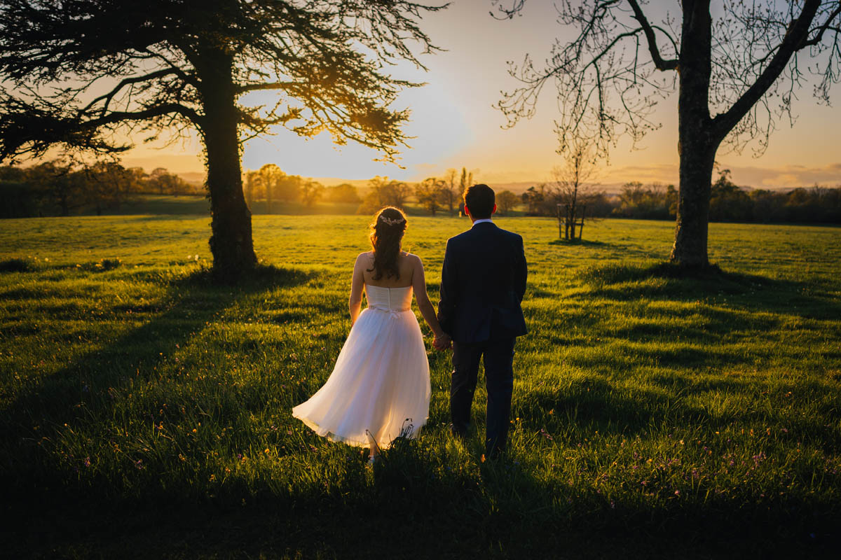 bride and groom walk towards the sunset amidst the trees at rockbeare manor