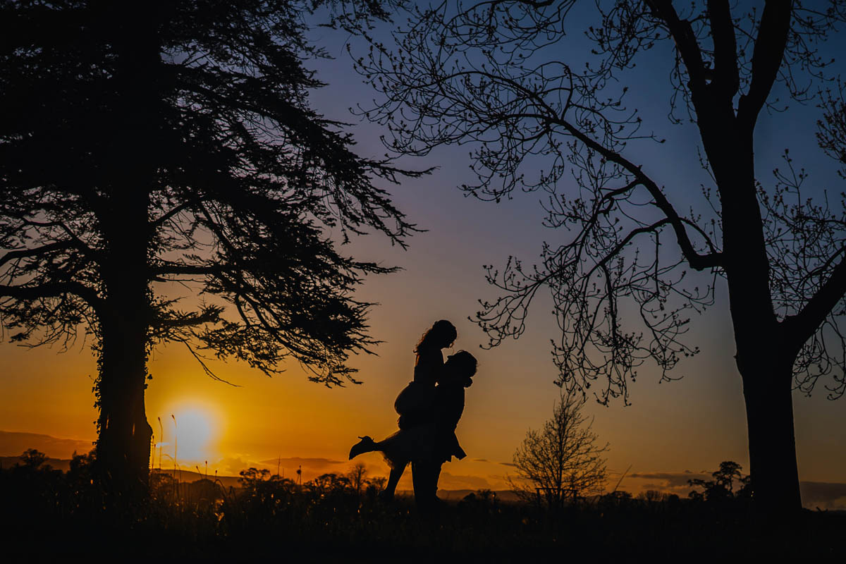 a silhouette photograph of the groom lifting his bride up. two trees create silhouettes too.