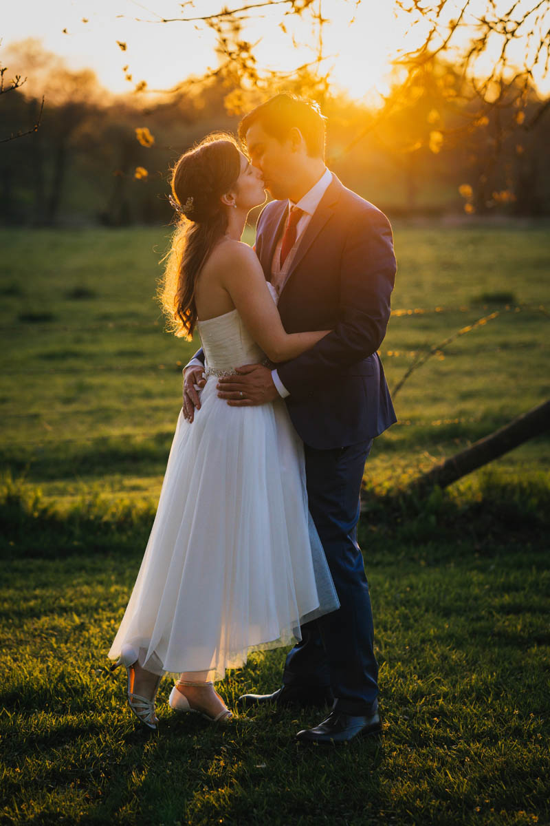 the newlyweds kiss with their arms around on another. behind them the sun glows as it sets in the evening sky