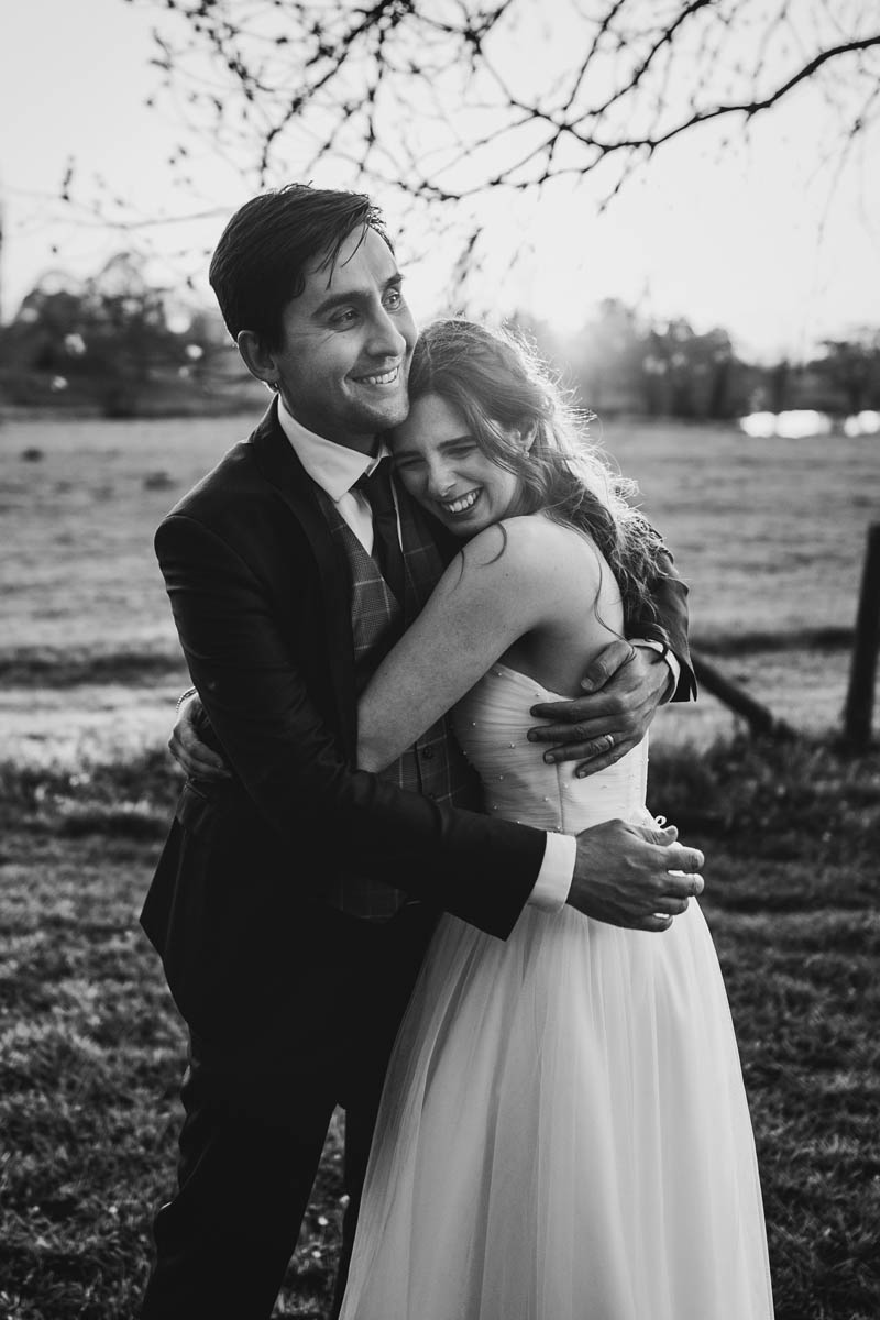 the groom hugs his bride as they both smile. in black and white