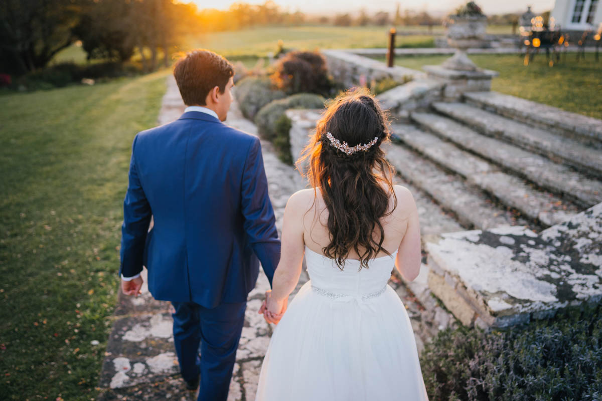 the golden hour light glows behind the newlyweds as they hold hands and walk towards the wedding venue