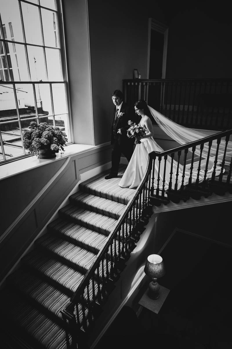 the father of the bride walks the bride down the staircase at rockbeare manor. a high contrast black and white photograph with strong highlights and shadows