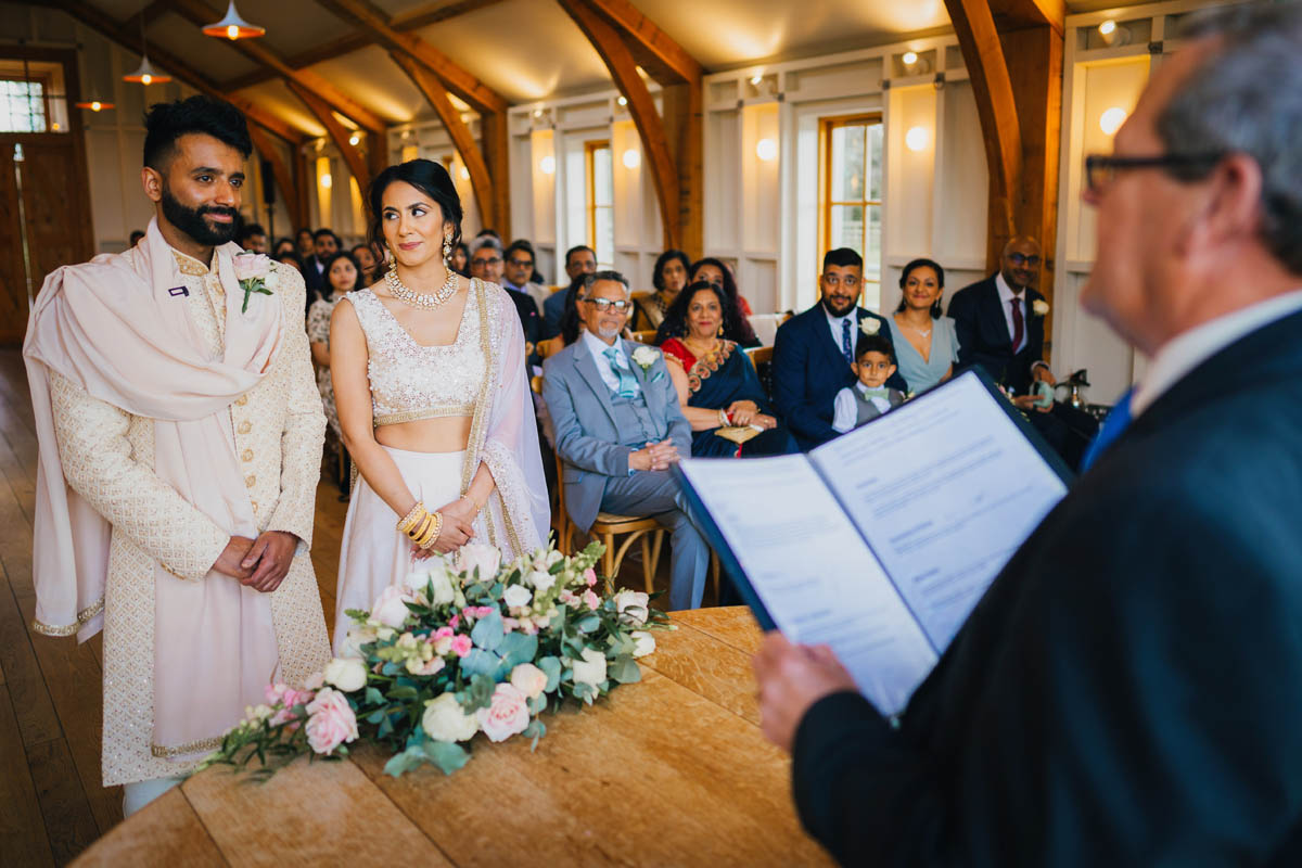 the marriage officiant reads from his book whilst the bride and groom smile at one another and wedding guests watch in the background