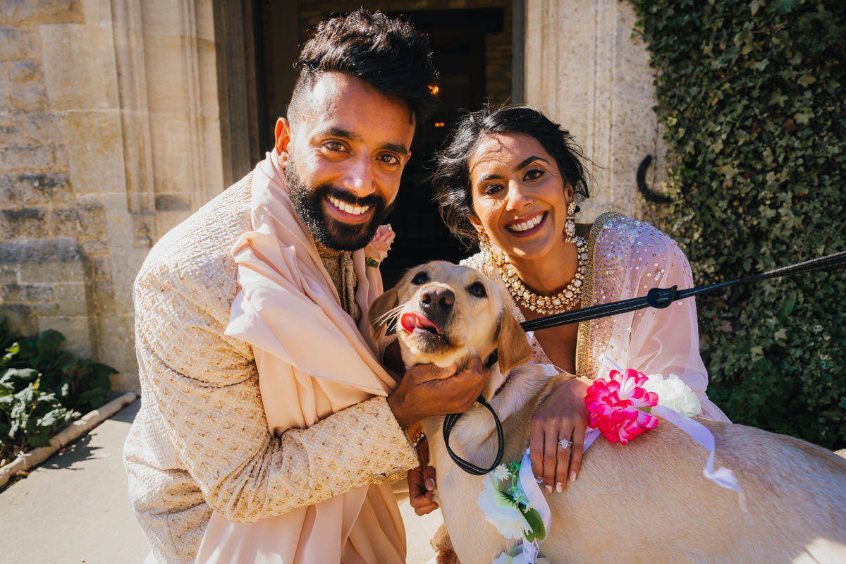 the bride and groom look into the camera and smile as they hug their golden Labrador who sticks her tongue out