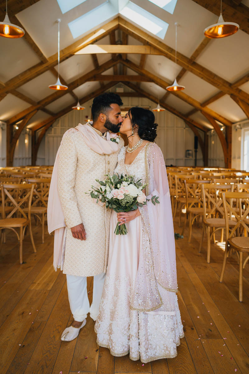 portrait photograph of both the newly-weds as they share a kiss. They are both dressed in beautiful cream and gold traditional Indian wedding outfits and the bride holds her wedding bouquet