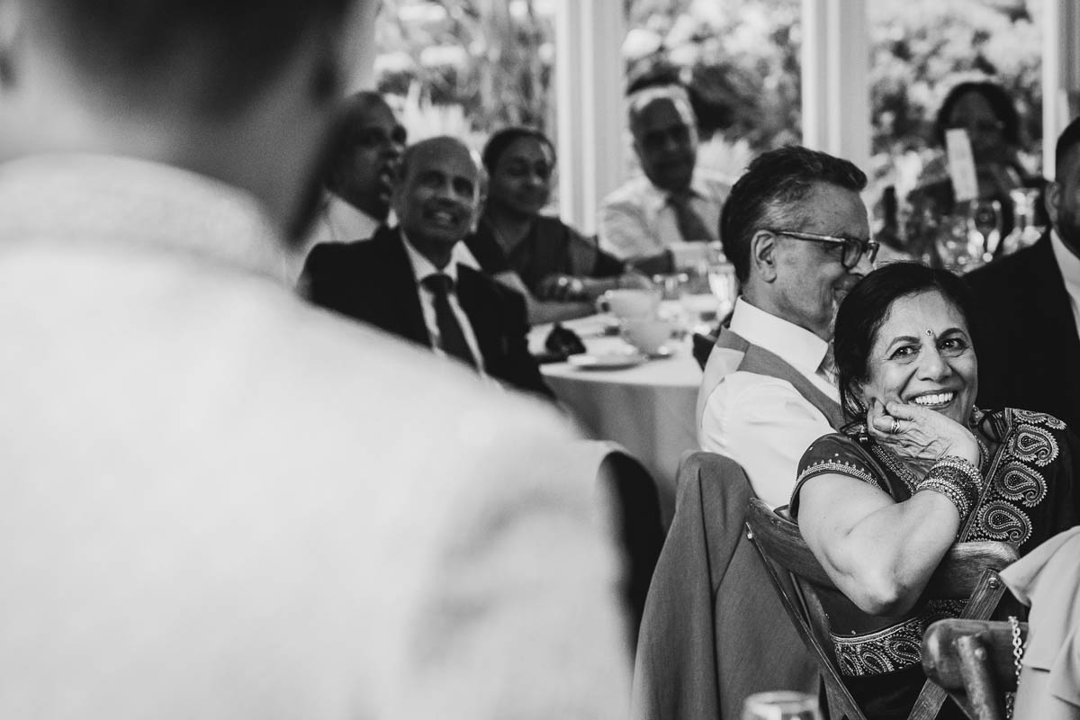the mother of the bride smiles as she listens to the groom's speech. Wedding guests are in the background, listening to the speech. IN black and white