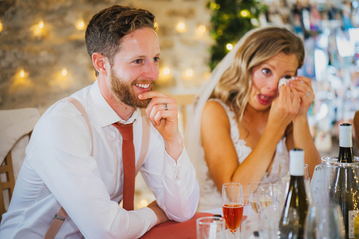 the groom smiles in the foreground as his new wife wipes away a tear in the background