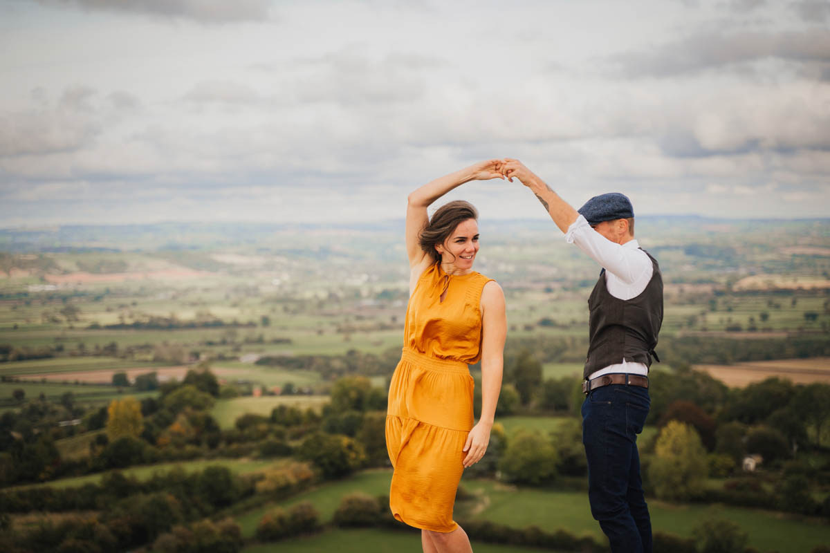 a woman in a yellow dress dances with a man in smart trousers, waist coat and flat cap with the Somerset countryside behind