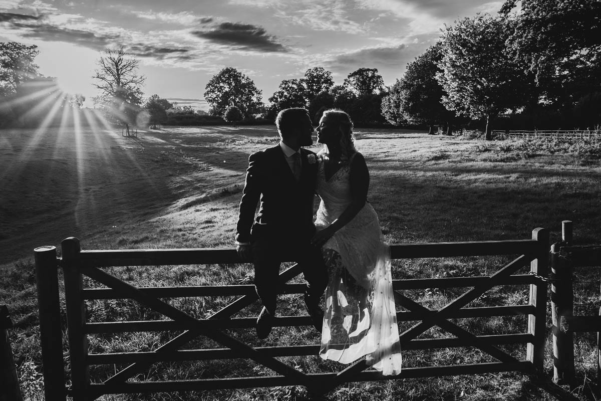 a black and white photograph of a newly married couple at sunset. the bride and groom sit on a gate