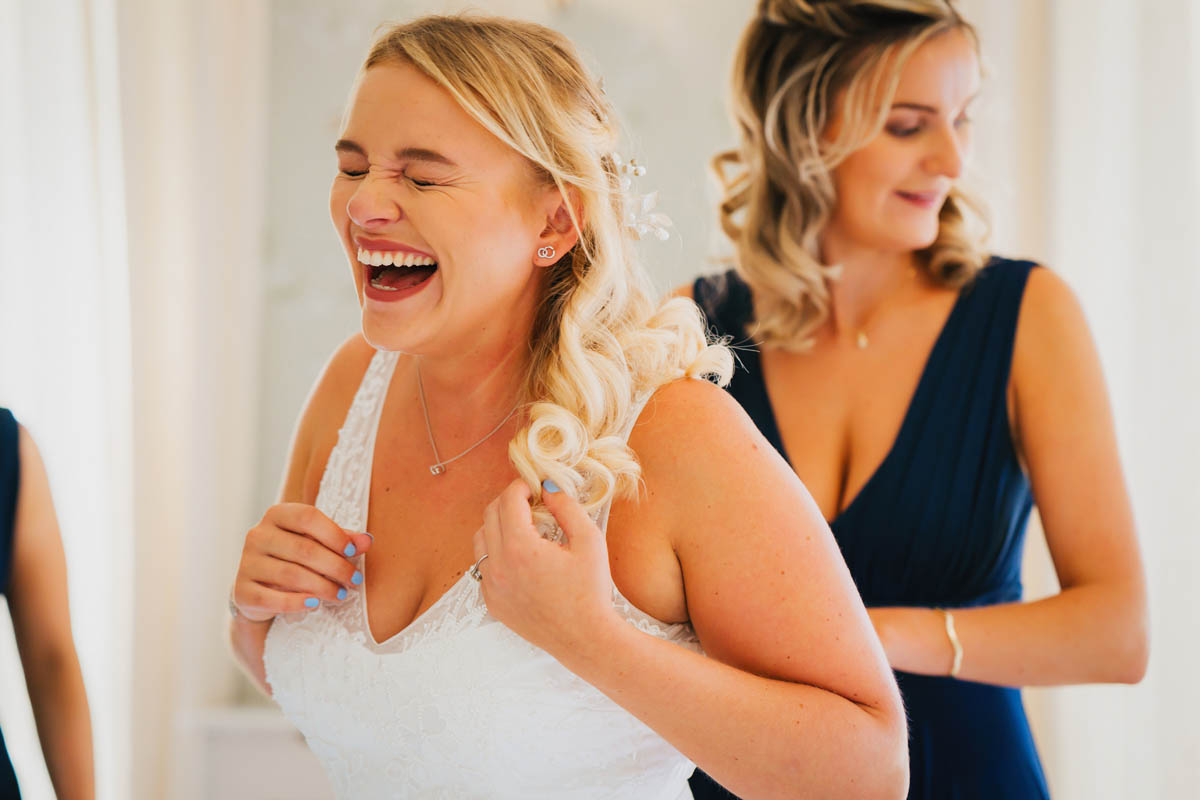 the bride laughs as her bridesmaid fixes her bridal gown