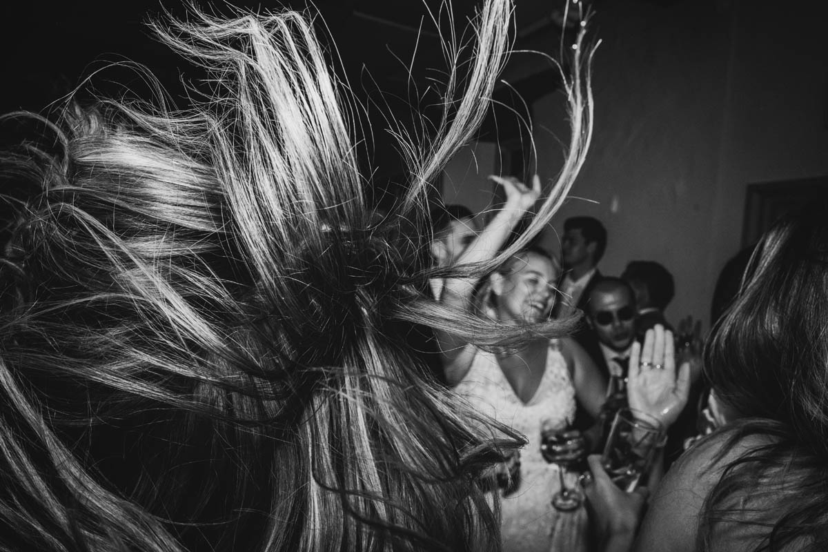 in the background the bride dances whilst the foreground is filled by a wedding guest throwing their hair back on the dance floor