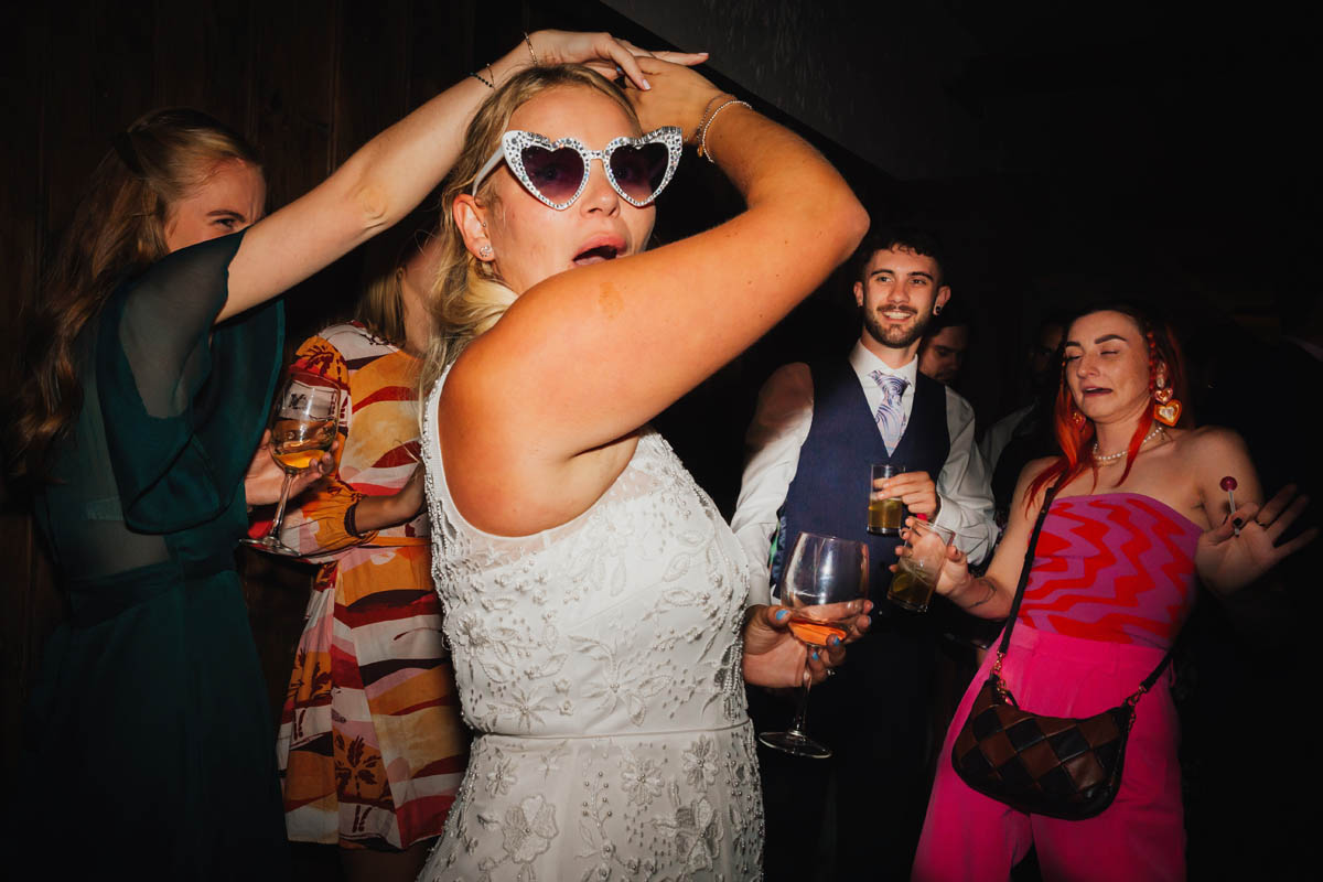 the bride spins around with her friend on the dance floor whilst wearing bit heart sunglasses