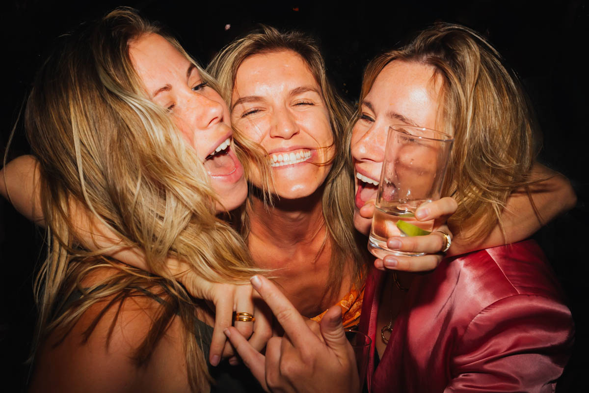 three white women with blonde hair smile and hold one another on the dance floor