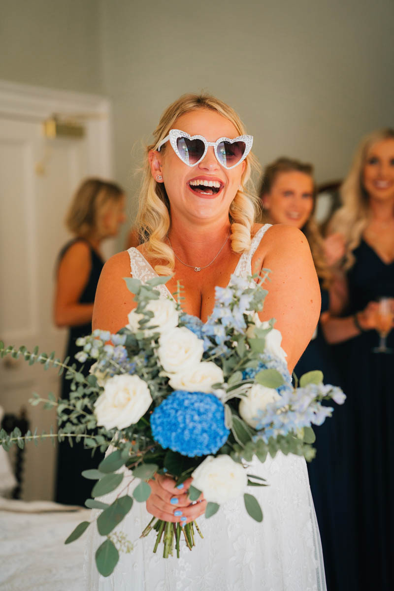 the bride poses with her bridal bouquet and wearing large silver love heart glasses