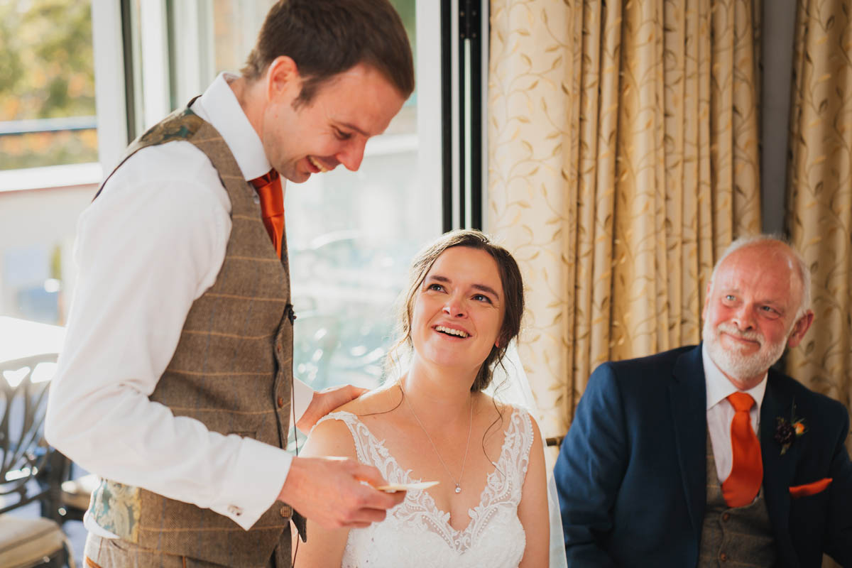 the groom deliver his speech whilst the bride and father of the bride watch and smile. The groom lovingly touches his bride's shoulder