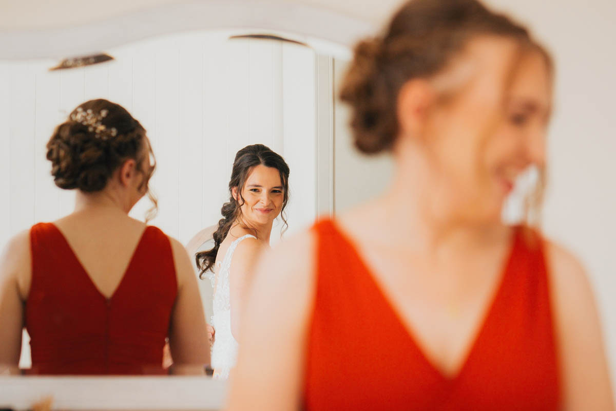 the bride smiles at her reflection in the mirror whilst the bridesmaids, wearing burnt orange dresses, look on