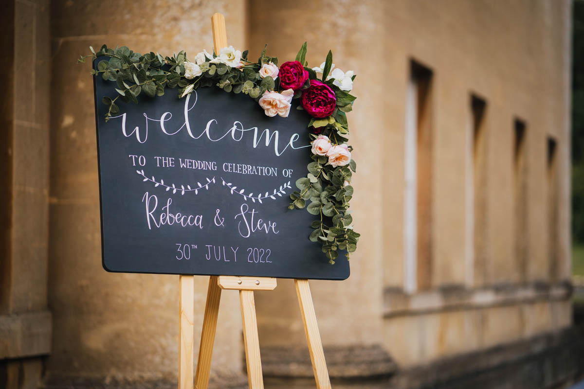 a welcome sign to the wedding with a bath stone wall behind