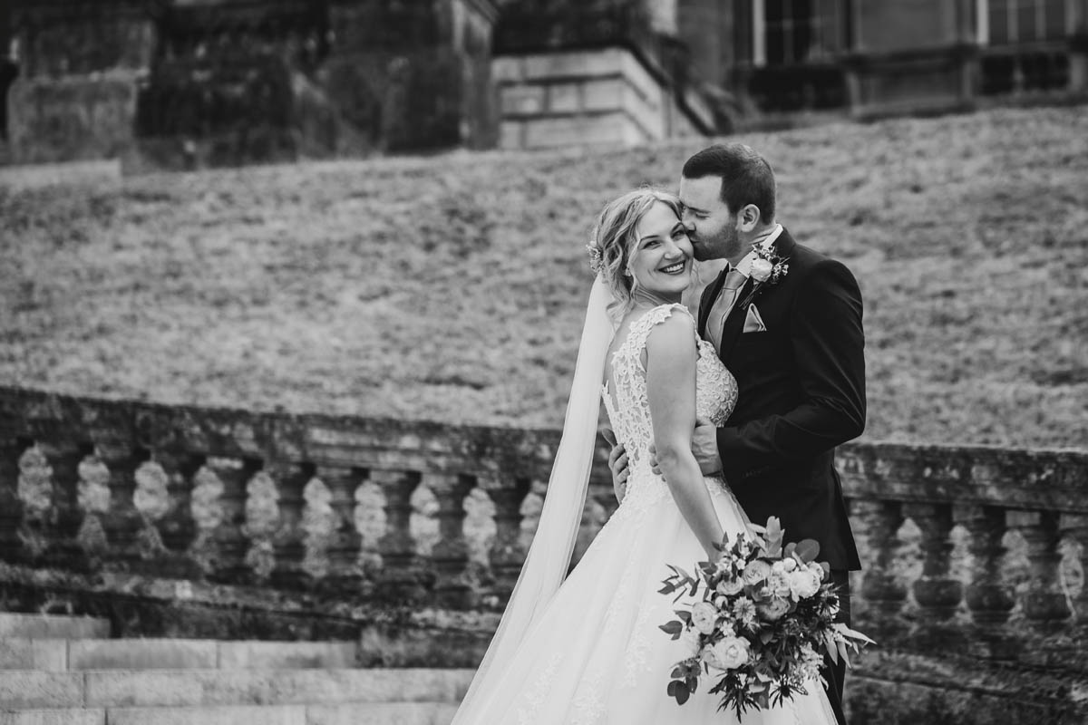 black and white image of the bride looking over her shoulder in her bridal gown with the groom holding her waist and kissing her cheek