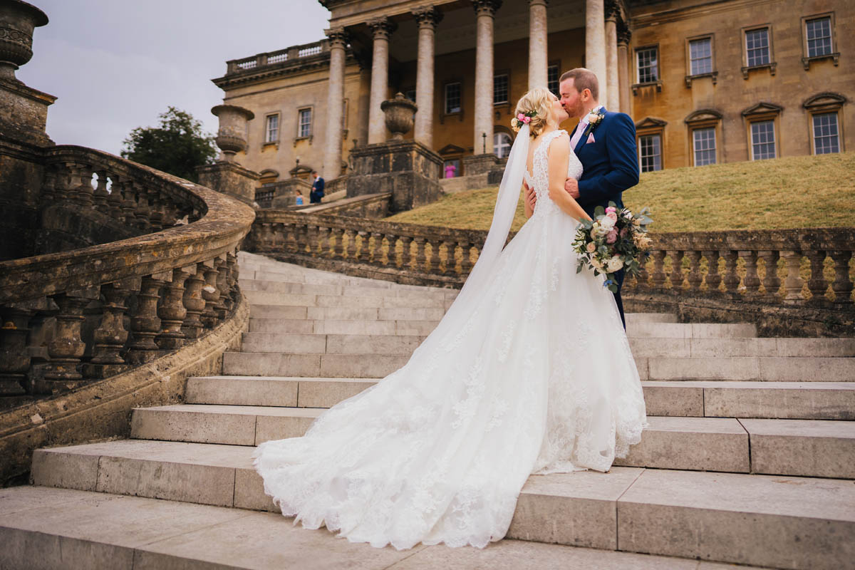 A man kisses his new wife in their bridal attire on the steps in front of prior park