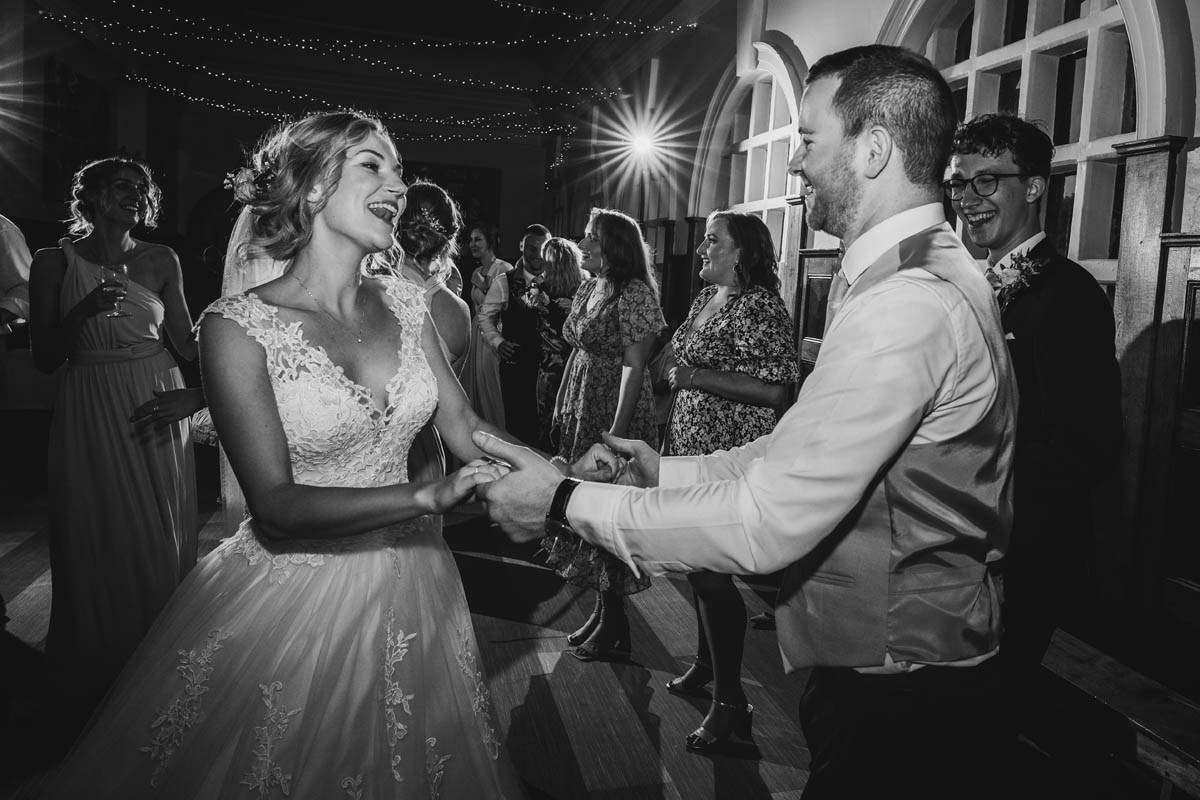 black and white image of the bride and groom dancing. Wedding guests join them behind. an off-camera flash fires into the camera