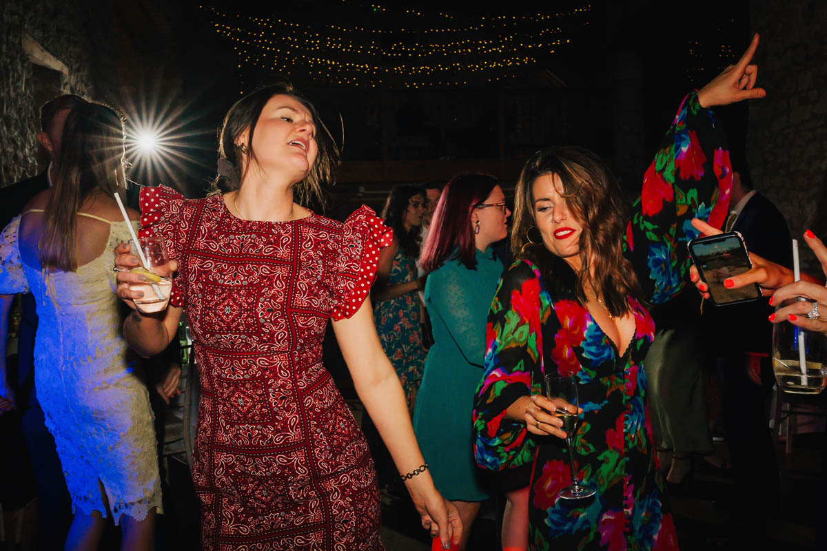 two women fling their arms into their air as they hold drinks on the dance floor at priston mill