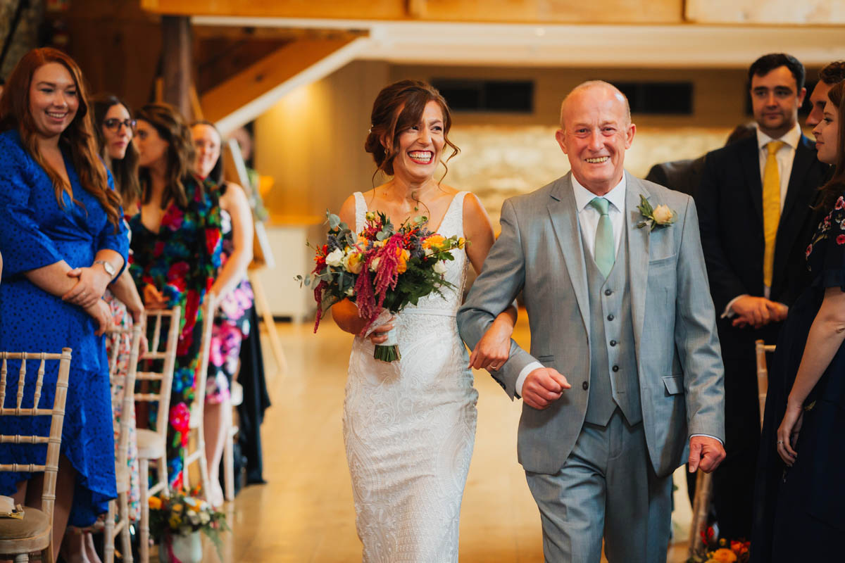 the father of the bride walks his smiley daughter down the aisle while happy wedding guests look on