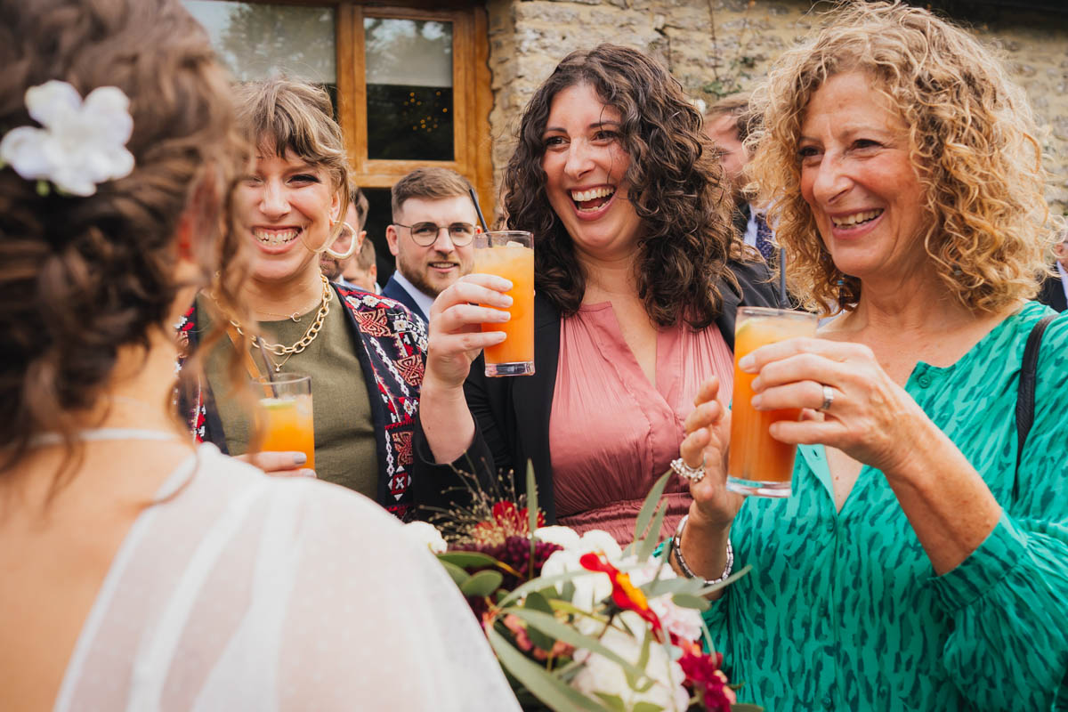 wedding guests smile as they hold cocktails and talk to the bride