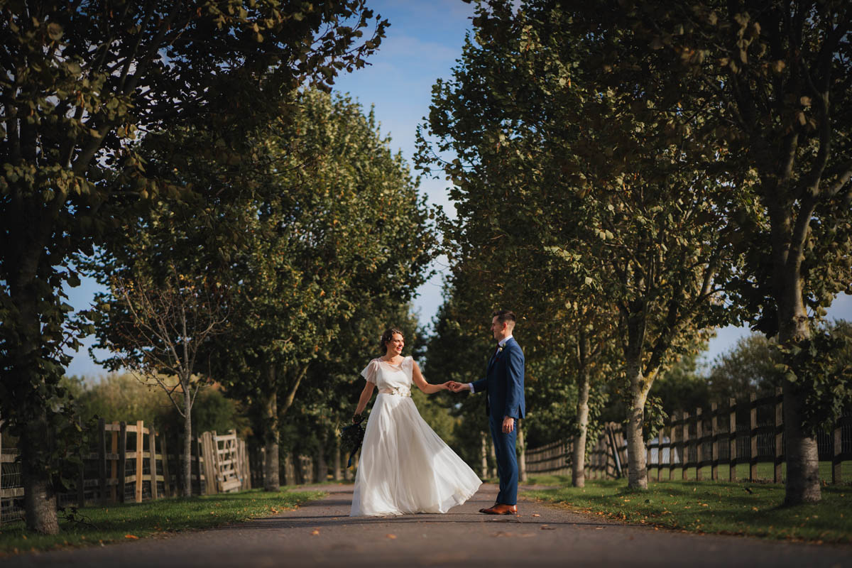 bride and groom walk down the autumnal drive with green trees lining the drive and autumn leaves on the floor