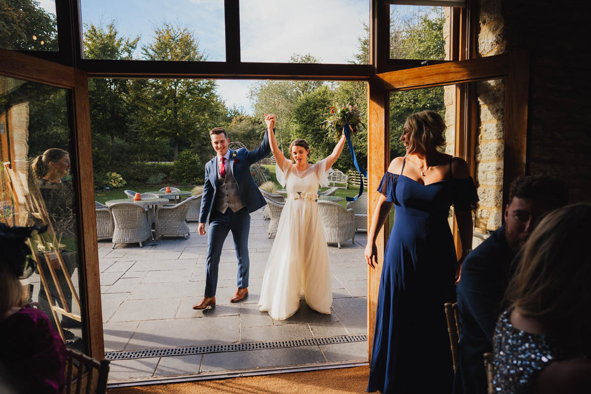 the foreground is dark and in the background the bride and groom enter the wedding breakfast room from outside, their arms in the air as they hold hands