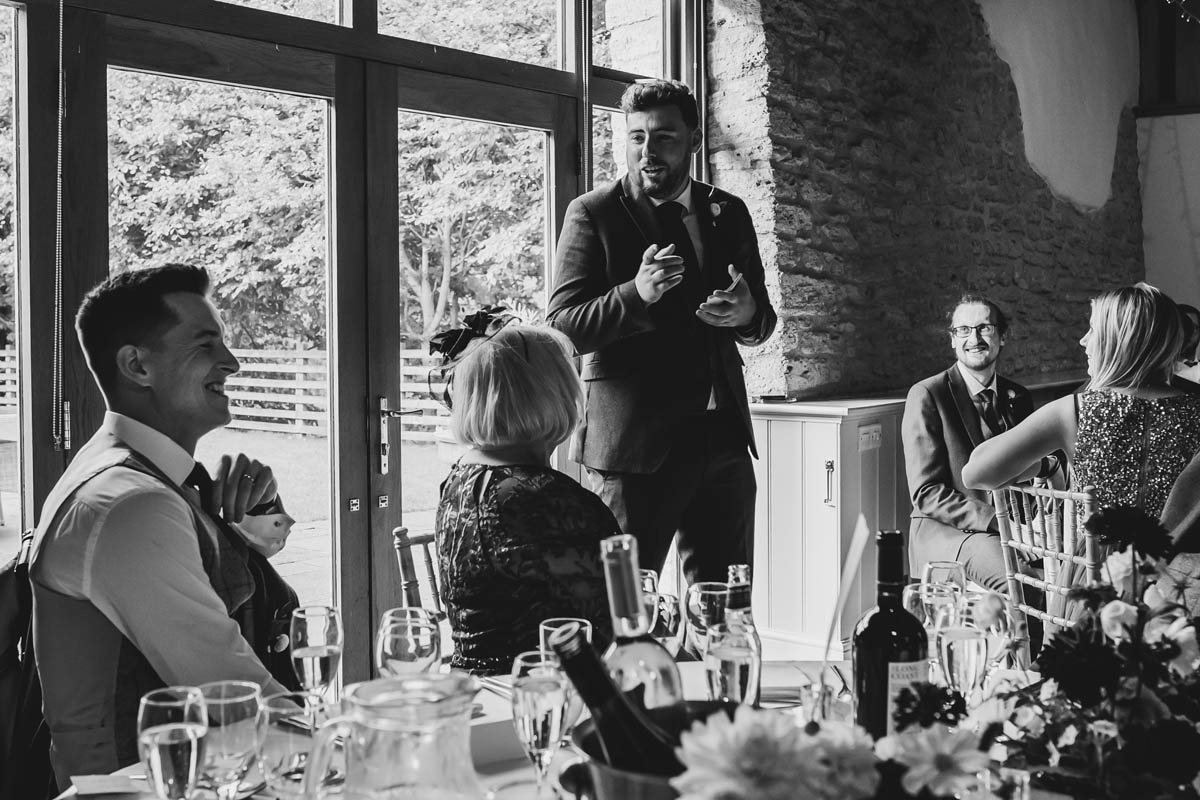 black and white photograph of the best man giving his speech, the groom and guests laugh