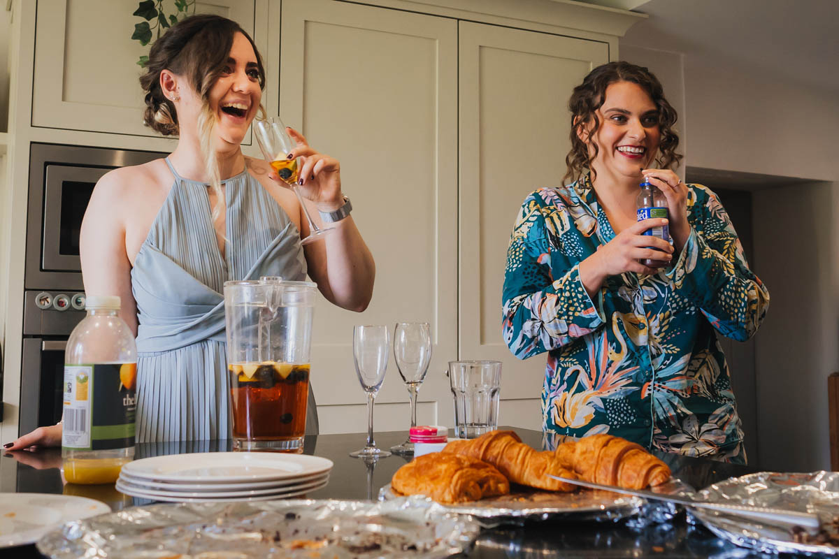 the bride in her pyjamas and a bridesmaid laugh as they drink champagne