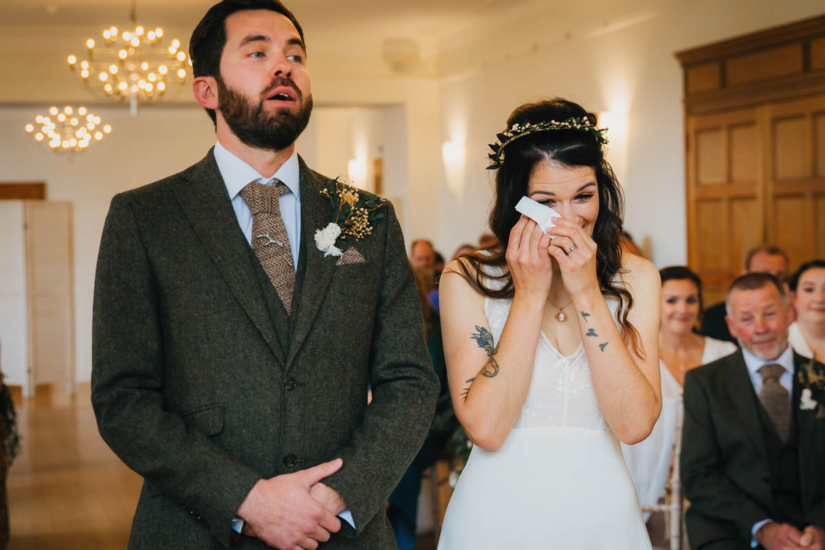 the groom looks emotional as he chokes back tears and his wife wipes away a tear during their wedding ceremony, wedding guests watch from behind