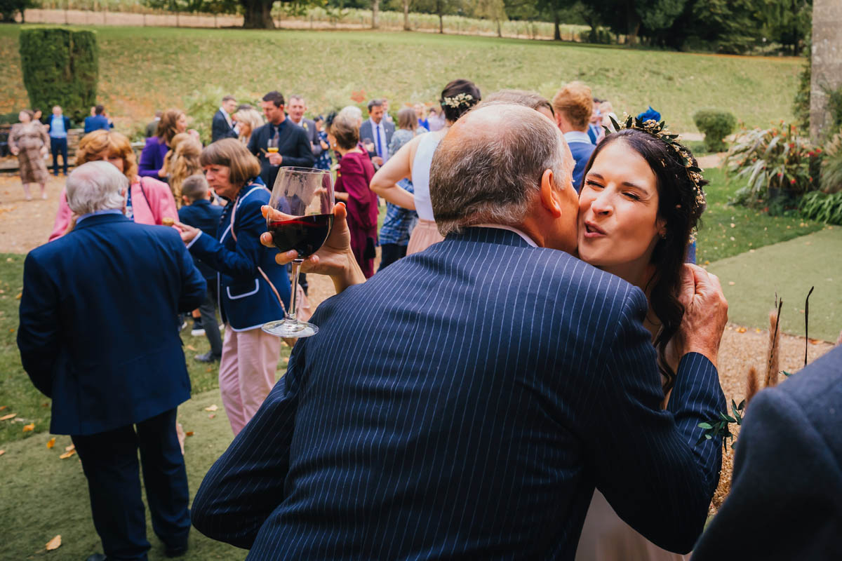a bride kissing a wedding guest whilst the other wedding guests enjoy the drinks reception behind