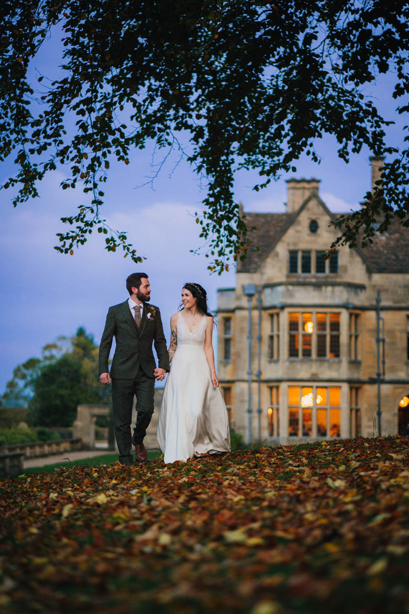 a bride and groom look at one another and smile as they walk away from the lit up wedding venue behind, Autumn leaves carpet the floor