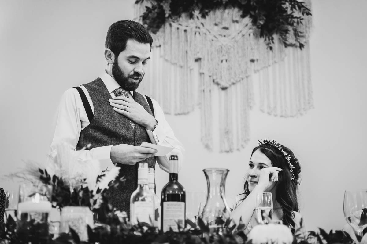 black and white photograph of the groom standing, delivering his speech whilst his new wife wipes away a tear next to him