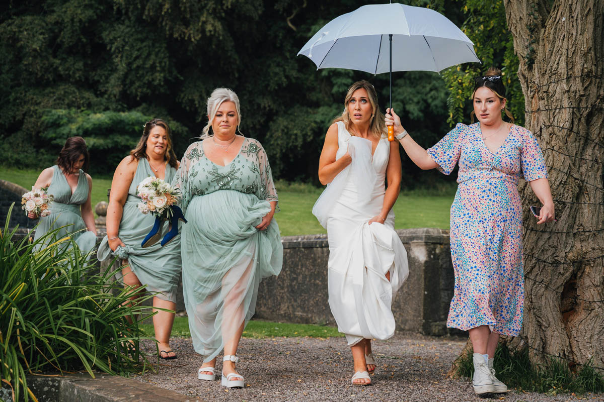 the bride looks concerned as venue staff hold an umbrella above her as she walks through barley wood gardens towards her wedding ceremony