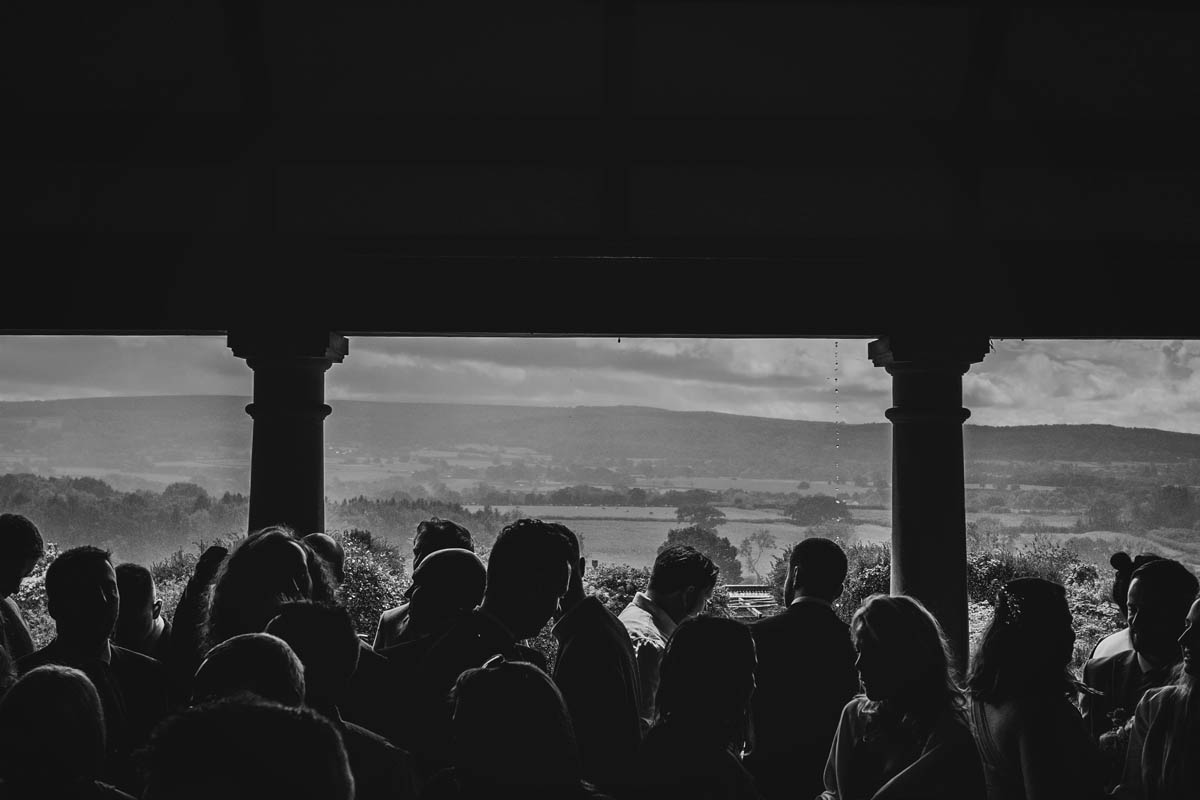 black and white photograph of wedding guests crowded into the summer house at barley wood while rain pours in the landscape behind them