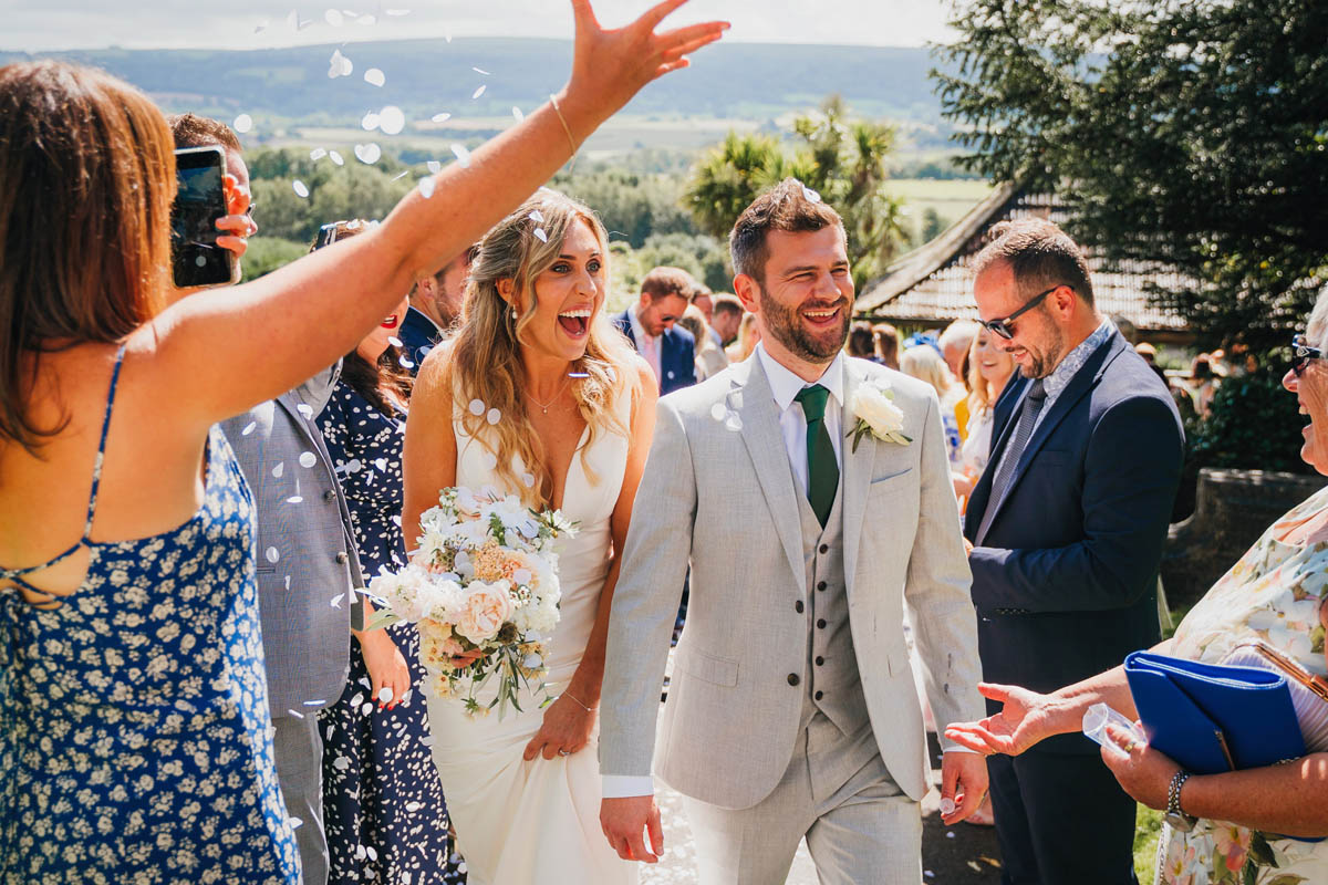 wedding guests throw confetti over a happy bride and groom with mendip hills in the background