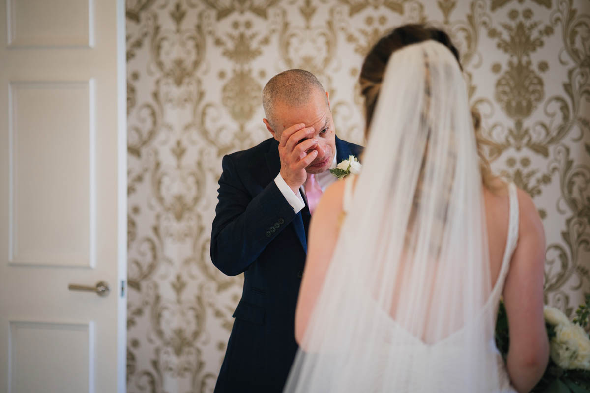 the father of the bride wipes a tear as he sees his daughter for the first time in her bridal dress