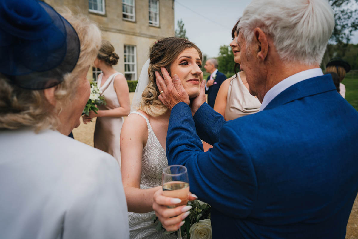 the bride's grandfather cups her face as she looks emotional whilst holding a glass of champagne
