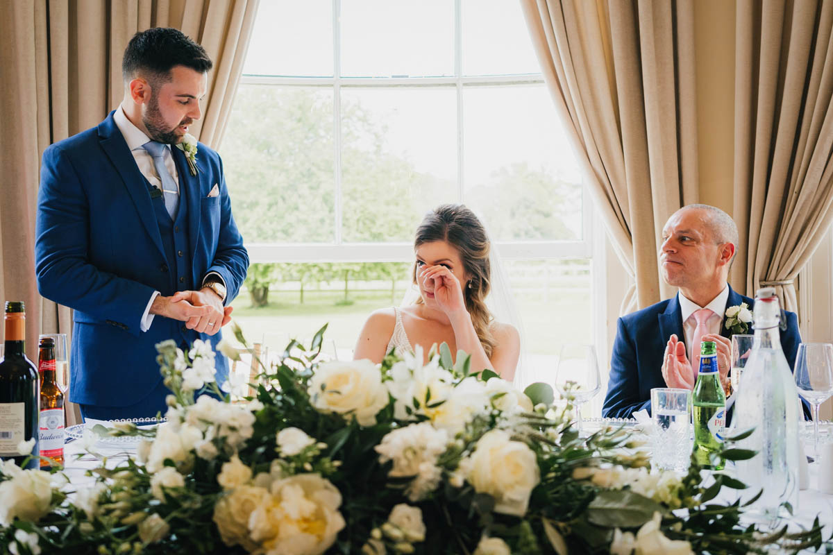 the groom delivering his speech whilst the bride shares a tear and the father of the bride looks on proudly