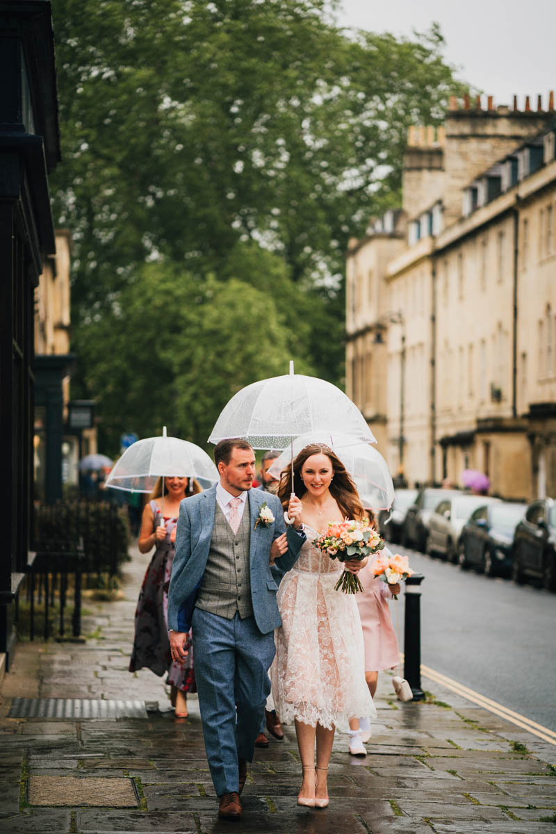 bride and groom holding an umbrella in the rain and walking through Bath