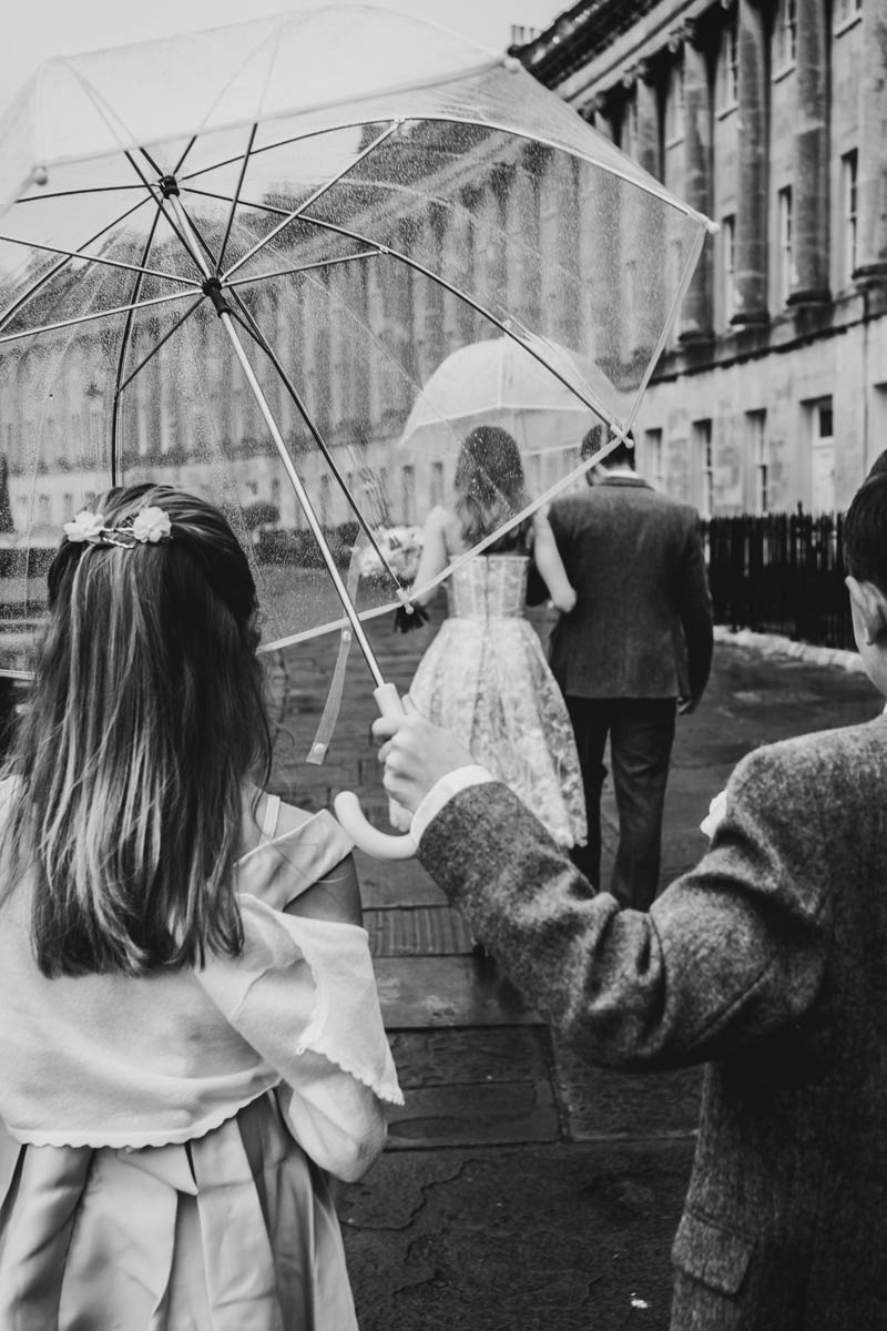 the bride, groom and their wedding guests walk in the rain through the royal crescent