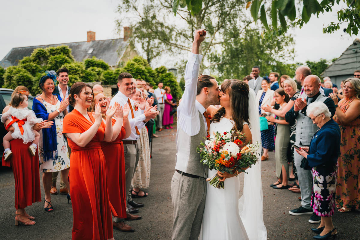 the groom throws his fist in the air and kisses his wife as guests clap in the background