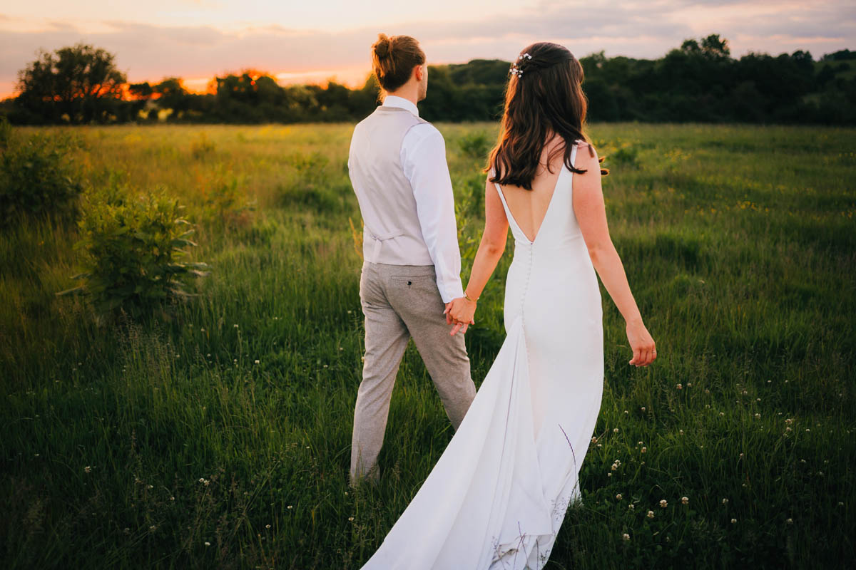 the groom leads his wife through a field at golden hour