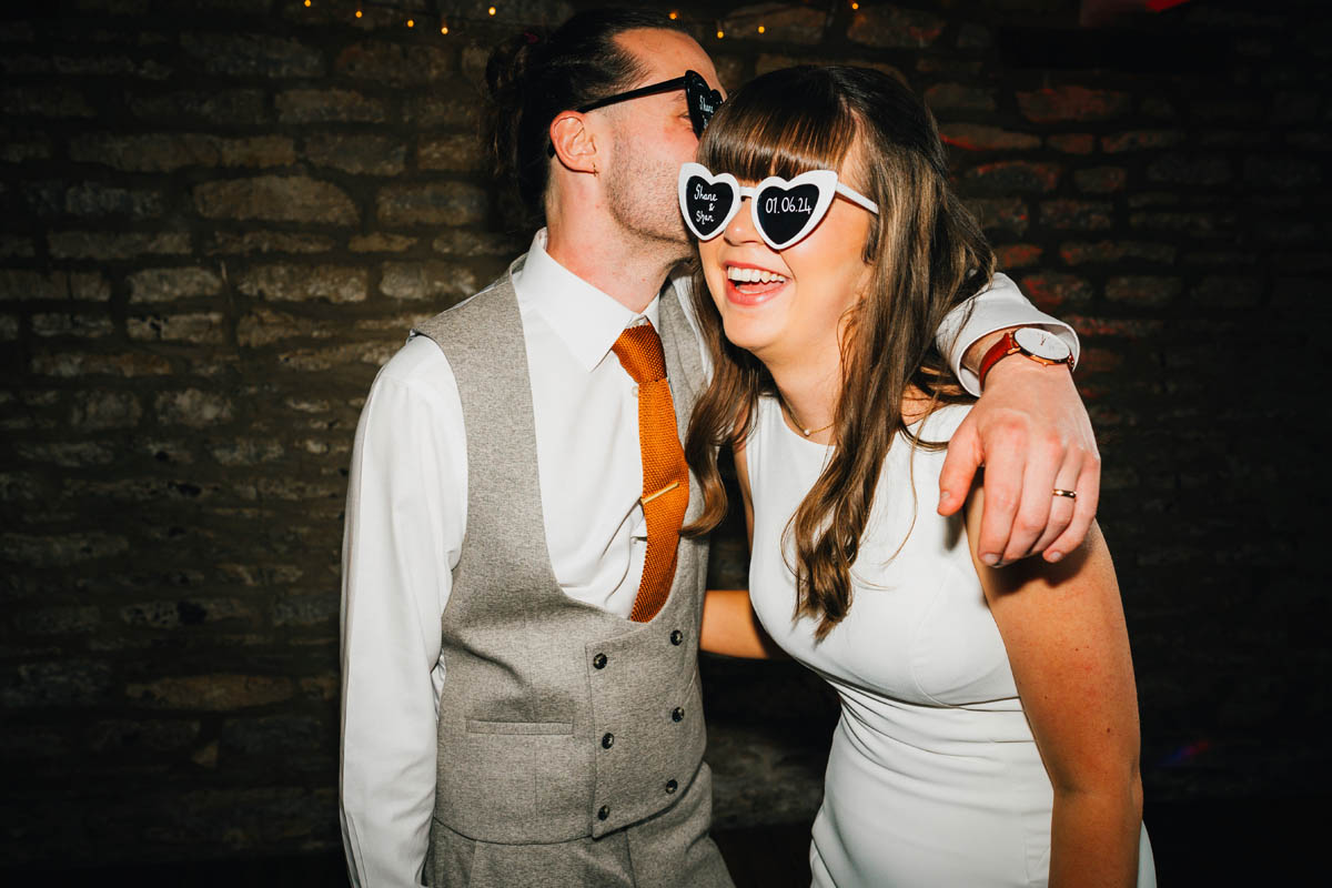 the groom kisses his wife who is wearing heart sunglasses with their wing details written on